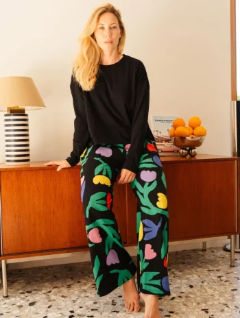 Mujer sentada descalza sobre un aparador, con jersey negro y pantalón floral multicolor